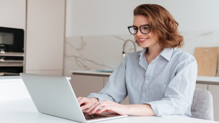 Mulher sorridente elegante de óculos e camisa listrada usando computador portátil enquanto está sentado à mesa de trabalho